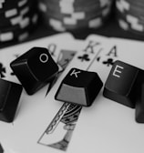 A series of playing cards and poker chips are displayed. The cards, including a ten, jack, queen, king, and ace of clubs, are spread out with black keyboard keys arranged to spell the word 'POKER'. The image is in black and white, giving it a classic, stylish look.