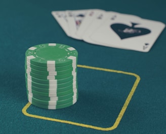 A stack of green poker chips is positioned on a green felt table within a yellow rectangular outline. In the background, there is a fanned set of playing cards, including an ace of spades visible.