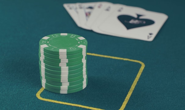 A stack of green poker chips is positioned on a green felt table within a yellow rectangular outline. In the background, there is a fanned set of playing cards, including an ace of spades visible.