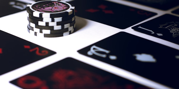 A stack of poker chips with a distinctive black and white pattern is placed on a table covered with several playing cards. The cards are dark with red and white symbols, creating a bold contrast against the lighter surface.