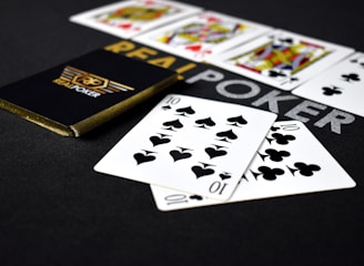 A deck of playing cards labeled 'Real Poker' rests on a dark surface. Two cards, the ten of spades and ten of clubs, are prominently displayed in the foreground. A row of face cards, including kings and queens from different suits, is lined up in the background.