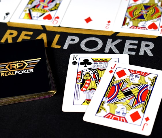 Poker-themed setup featuring a deck of cards on a table with the words 'REAL POKER' visible. Two cards, the King of Clubs and Jack of Diamonds, are prominently displayed. The design is vibrant, with detailed illustrations on the cards.