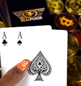 A hand holding two playing cards, an Ace of Clubs and an Ace of Spades, with a backdrop of gold Bitcoin tokens. The hand has an orange fingernail with a white Bitcoin symbol on it. The background features a stack of poker chips with a logo that reads 'Real Poker'.