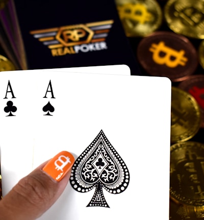A hand holding two playing cards, an Ace of Clubs and an Ace of Spades, with a backdrop of gold Bitcoin tokens. The hand has an orange fingernail with a white Bitcoin symbol on it. The background features a stack of poker chips with a logo that reads 'Real Poker'.