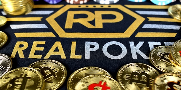 Stacks of poker chips in various colors are arranged in the background, while in the foreground, there are numerous golden coins featuring the Bitcoin symbol. The text 'REALPOKER' is prominently displayed in gold and silver lettering on a dark background.