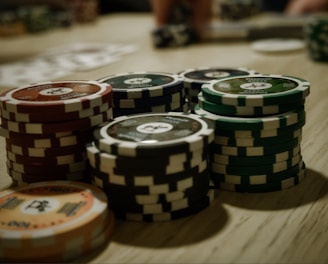 Stacks of poker chips are arranged on a wooden table, with various denominations and colors such as red, green, and black. Some playing cards are blurred in the background, indicating a game setting.