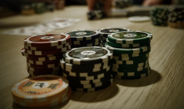 Stacks of poker chips are arranged on a wooden table, with various denominations and colors such as red, green, and black. Some playing cards are blurred in the background, indicating a game setting.