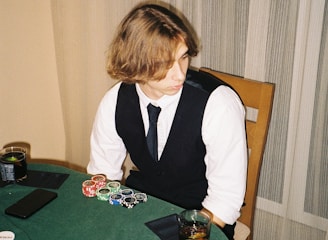 A person with shoulder-length hair is sitting at a poker table. They are wearing a white shirt, black vest, and tie, with an array of poker chips in front of them. There are two glasses with drinks on the table, and a smartphone is placed nearby. The background consists of vertical striped curtains.