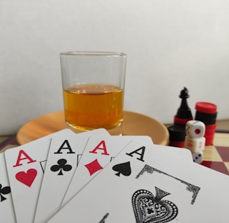A hand of playing cards featuring four aces and a three of clubs is displayed over a checkered board. A glass of amber liquid rests on an orange saucer in the background. There are stacked poker chips, dice, and a black chess piece nearby.