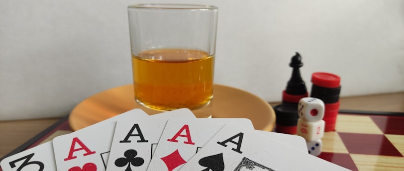 A hand of playing cards featuring four aces and a three of clubs is displayed over a checkered board. A glass of amber liquid rests on an orange saucer in the background. There are stacked poker chips, dice, and a black chess piece nearby.